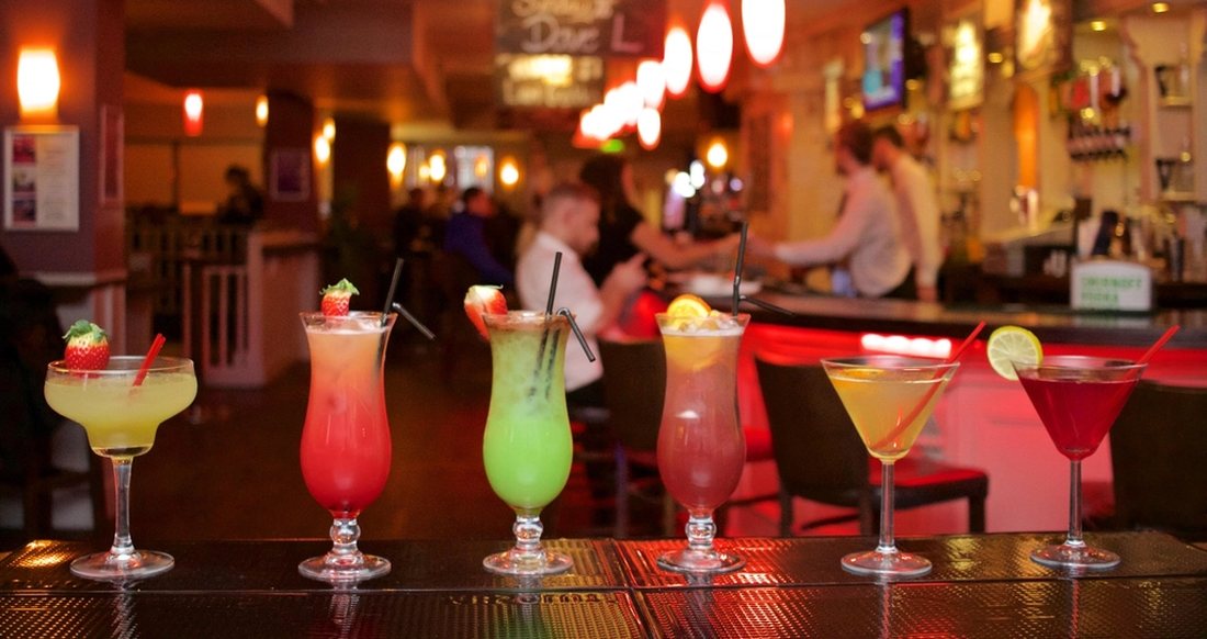 A row of cocktails on a lit bar with staff working behind it